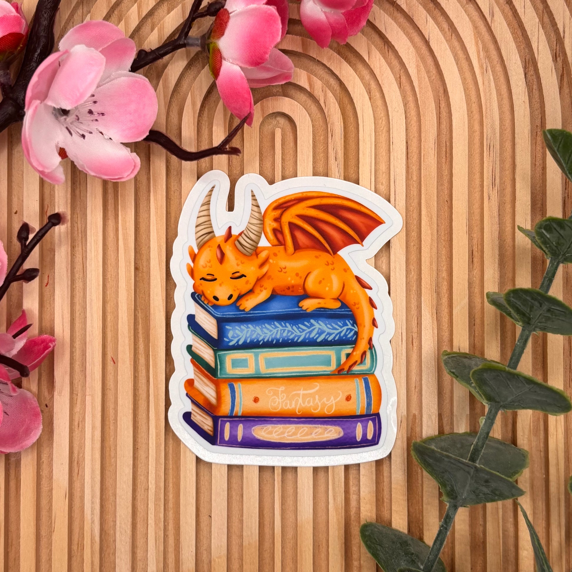 Die-cut sticker van oranje draak slapend op stapel boeken.